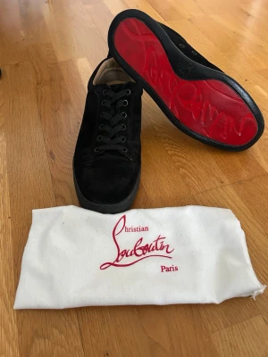 Svarta sneakers från Christian Louboutin - Snygga svarta sneakers i mocka från Christian Louboutin med den ikoniska röda sulan. Skorna har svarta skosnören, diskret design och beige innerfoder. Perfekta för dig som vill ha en exklusiv och stilren look.