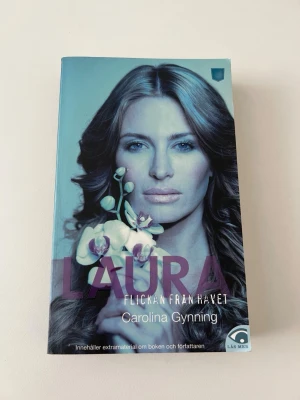 Laura - Flickan från havet - Romanen 'Laura - Flickan från havet' av Carolina Gynning är en pocketbok med ett blåtonat omslag och ett porträtt av en kvinna med en orkidé. Boken innehåller extramaterial om författaren och boken. Perfekt för dig som gillar spännande och psykologiska berättelser.