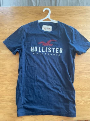 Marinblå Hollister t-shirt - Snygg marinblå t-shirt från Hollister med stort tryck på bröstet i vitt och vinrött. Klassisk rund hals och korta ärmar. Perfekt till jeans eller shorts för en avslappnad stil.