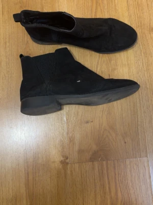 Svarta Chelsea boots i mocka - Säljer ett par klassiska svarta Chelsea boots i mocka med elastiska paneler på sidorna och låg klack. Skorna har rundad tå och dragflik bak för enkel påtagning. Perfekta för dig som gillar stilrena och tidlösa skor.