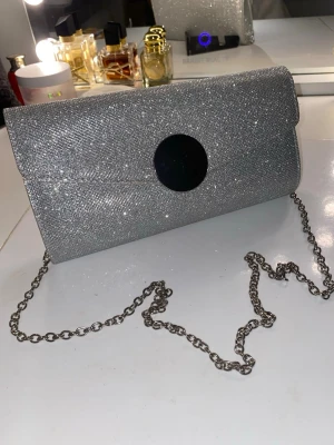 Glittrig silver kuvertväska med kedja - Använd en gång. Snygg kuvertväska i glittrigt silver med en rund svart detalj på locket och en lång silverfärgad kedja. Väskan har ett innerfack och stängs med magnetlås. Perfekt för att lyfta din outfit med extra bling.