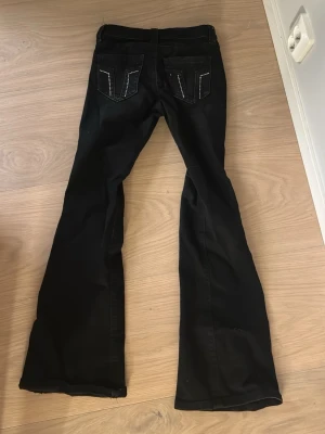 Svarta bootcut jeans med detaljer - Säljer ett par svarta bootcut jeans med snygga kontrastsömmar och dekorativa fickor bak. Jeansen har utsvängda ben och coola detaljer på bakfickorna. Perfekta för dig som gillar en retroinspirerad look och vill sticka ut lite extra.