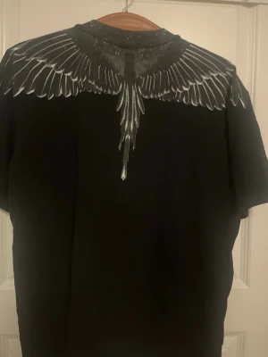 Svart t-shirt med vingtryck Marcelo Burlon - Säljer en svart t-shirt från Marcelo Burlon County of Milan med ikoniskt vingtryck i vitt och grått över axlar och rygg. Tillverkad i 100% bomull och har en avslappnad passform. Perfekt för dig som gillar streetwear och unika prints.