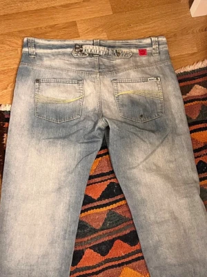 Ljusblå raka jeans med detaljer - Säljer ett par ljusblå jeans med raka ben och slitna tvättade detaljer. Jeansen har två bakfickor med gul söm och knappdetaljer samt en justerbar rem i midjan. Materialet är klassisk denim i bomull. Midjemåttet tvärs över är 44.