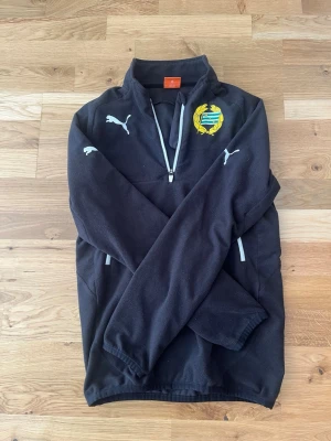 Svart fleecejacka Hammarby Puma - Svart fleecejacka från Puma med Hammarby IF:s klubbmärke på bröstet. Jackan har halv dragkedja framtill, ståkrage och vita Puma-loggor på bröst och ärm. Perfekt för träning eller att ha på väg till match.