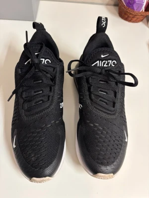 Nike Air 270 svarta sneakers - Svarta Nike Air 270 sneakers med vit sula och tydlig logga på sidan och plösen. Ovandelen är i meshmaterial för bra ventilation och skon har en synlig luftdämpning i hälen. Klassisk snörning och sportig design som passar till många stilar.