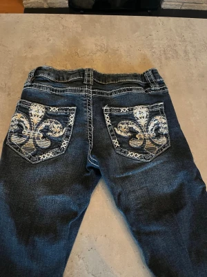 Mörkblå jeans med broderade fickor - Säljer ett par mörkblå jeans med utsvängda ben och snygga broderade fickor bak med fleur-de-lis-motiv i vitt och beige. Jeansen har kontrastsömmar och en cool tvättad look. Perfekta för dig som vill sticka ut med detaljer!