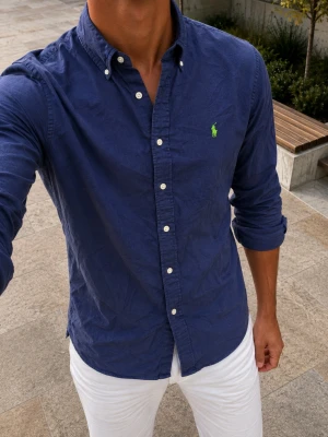 Ralph lauren skjorta mörkblå - Snygg mörkblå skjorta med klassisk krage och vita knappar. Skjortan har lång ärm och en liten grön broderad logga på bröstet. Perfekt till jeans eller chinos för en clean och stilren look.