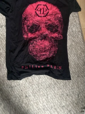Svart t-shirt med röd dödskalle Philipp Plein - Svart t-shirt från Philipp Plein med stor grafisk dödskalle i rött framtill och logga tryckt under motivet. Klassisk passform och rund halsringning. Perfekt för dig som gillar edgy streetstyle och vill sticka ut.