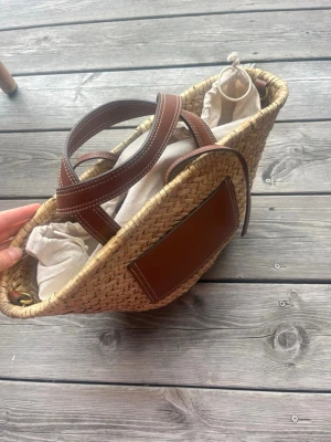 Stråväska med bruna detaljer - Snygg shoppingväska i flätad strå med bruna läderhandtag och matchande läderdetalj på framsidan. Väskan har en tygpåse inuti som kan dras åt med snöre. Perfekt för dig som vill ha en trendig och rymlig väska till sommaren.