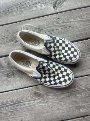 Vans slip-on checkerboard sneakers - Klassiska Vans slip-on sneakers med svartvitt checkerboard-mönster. Skorna har vit sula och elastiska paneler på sidorna för enkel påtagning. Tillverkade i canvasmaterial och har Vans-logga bak på hälen.