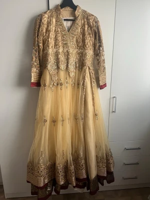 Champagnefärgad långklänning med broderi - Elegant långklänning i champagnefärg med detaljerat guldigt broderi och paljetter. Klänningen har långärmade spetsärmar, hög krage och vackra volangdetaljer. Kjolen är i flera lager tyll med broderad kant och inslag av vinrött vid ärmslut och nederkant.