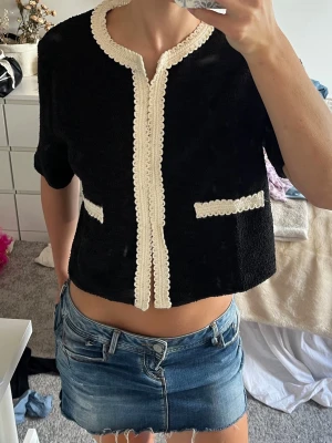 Svart kort kofta med spetsdetaljer - Kort svart kofta med vita spetsdetaljer längs kanterna och vid de två fickorna framtill. Koftan har rund halsringning och korta ärmar. Perfekt för en chic och trendig look.