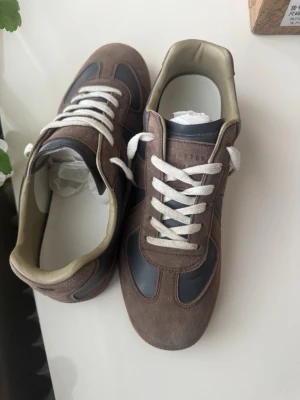 Margiela gats skor - Säljer ett par snygga sneakers i brunt mocka och grått skinn med vita snören. Skorna har en klassisk retrodesign med beige insida och gummisula. Perfekta för dig som gillar stilrena och tidlösa skor. Och som behöver skor till skolan.