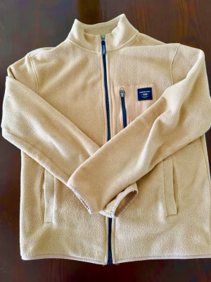 Lexington Beige fleecejacka  - Mysig beige fleecejacka från Lexington med hel dragkedja framtill och hög krage. Jackan har en bröstficka med dragkedja och Lexington-logga, samt två sidofickor. Perfekt för lager-på-lager och chill dagar.