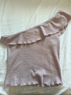 Ljusrosa oneshoulder topp från H&M - Säljer en ljusrosa oneshoulder topp från H&M med volangdetalj längs axeln i storlek XS.