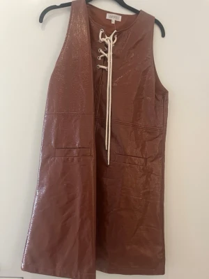 Brun ärmlös klänning med snörning från Ghospell - Snygg brun kortklänning i glansigt material med snörning framtill och två fickor. Ärmlös design och rak passform ger en cool retrovibe. Perfekt för dig som vill sticka ut med en unik look.