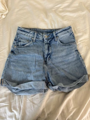 Blå denimshorts med upprullad kant - Säljer ett par klassiska blå jeansshorts i denim med hög midja och upprullade benslut. Shortsen har fem fickor och stängs med knapp och dragkedja. Perfekta till sommaren och enkla att matcha med allt.