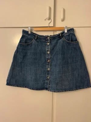 Blå jeanskjol med knappar framtill - Säljer en klassisk blå jeanskjol i kort modell med knappar hela vägen framtill. Kjolen har fickor fram och hällor för bälte, perfekt för en avslappnad och trendig look. Materialet är slitstarkt denim som passar till det mesta.
