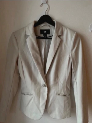 Beige kavaj från H&M - Stilren beige kavaj från H&M med klassisk krage och en knapp framtill. Kavajen har två fickor med lock och figurnära passform. Perfekt för en snygg och clean look.
