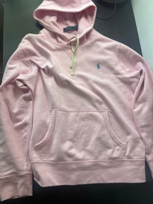 Ljusrosa hoodie från Polo Ralph Lauren - Säljer en ljusrosa hoodie från Polo Ralph Lauren med klassisk huva och vit dragsko. Tröjan har en känguruficka framtill och den ikoniska blå loggan broderad på bröstet. Tillverkad i mjukt bomullsmaterial som är riktigt skönt att ha på sig. Och jag köpte den för 2300kr.