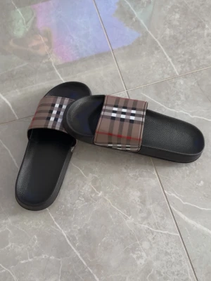 Svarta sandaler med rutigt band - Säljer ett par svarta sandaler med breda band i klassiskt beige, brunt och rött rutmönster. Sandalerna har en enkel slip-in design och en bekväm, lätt vadderad sula i syntetmaterial. Perfekta för sommaren och riktigt stilrena.