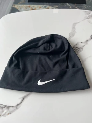 Svart mössa från Nike - Snygg svart mössa från Nike med klassisk vit logga framtill. Tillverkad i ett stretchigt och mjukt material som sitter skönt på huvudet. Perfekt för dig som vill ha en enkel och sportig stil under kyliga dagar.