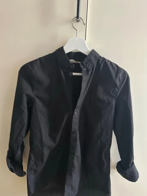 Svart långärmad skjorta - Stilren svart skjorta med lång ärm och klassisk krage. Skjortan har knappar framtill och är tillverkad i ett mjukt bomullstyg. Perfekt för dig som gillar en enkel och clean look.