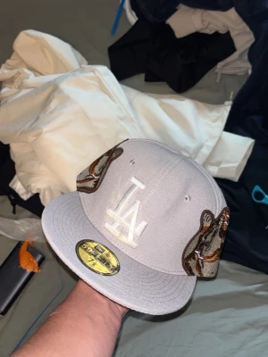 Grå LA keps med broderade ormar - Snygg grå keps med LA-logga framtill i vitt och broderade ormar på sidorna. Platt skärm och broderad text bak. Klassisk streetwear-stil, perfekt för dig som vill sticka ut med detaljer. Justerbar passform och cool design.