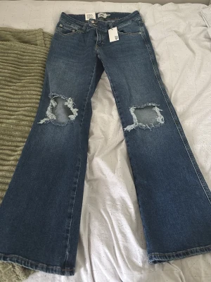 Blå bootcut jeans med slitna knän - Säljer ett par blå bootcut jeans med stora slitningar över båda knäna. Jeansen har klassisk femficksdesign och är gjorda i denim. Perfekta för dig som gillar en avslappnad och trendig stil.