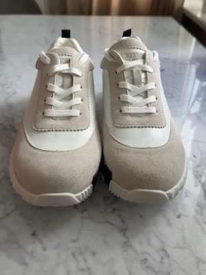 Vita och beige sneakers med mockadetaljer - Stilrena sneakers i vitt och beige med ovandel i slät syntet och mockadetaljer. Skorna har vita snören, räfflad sula och en svart detalj på sidan av sulan. Perfekta för en clean och modern look.