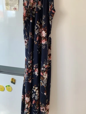 Mörkblå blommig långklänning - Snygg långklänning i mörkblått med blommigt mönster i rosa, vitt och beige. Klänningen är ärmlös och har en mjuk, flytande passform som ger en avslappnad men elegant look. Perfekt för sommarens alla tillfällen!