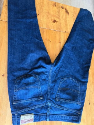 Mörkblå raka jeans - Säljer ett par mörkblå jeans med raka ben och klassiska bakfickor. Jeansen har en enkel design utan slitningar och är tillverkade i ett robust denimtyg. Perfekta för dig som gillar en tidlös och clean look. Mc Gordon 36/34