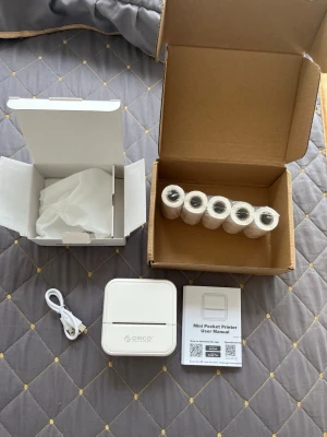 [ORICO] Mini pocket printer med papper - Säljer en vit, kompakt mini termisk skrivare från ORICO. Medföljer laddningskabel, användarmanual och flera rullar termopapper. Perfekt för att skriva ut små lappar, etiketter eller anteckningar direkt från mobilen. Enkel att ta med tack vare sin lilla storlek.