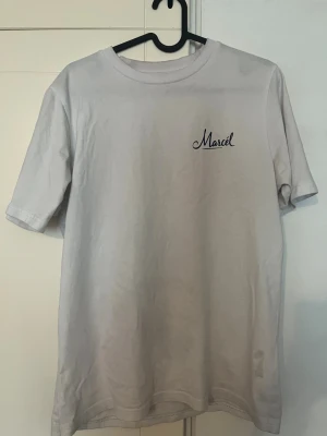 Vit T-Shirt från Leon Marcel - Säljer en vit t-shirt med trycket 'Marcel' på bröstet och ett stort motiv på ryggen med vinflaska, glas och båt. T-shirten har rund hals och korta ärmar. Perfekt för dig som gillar unika prints och stilrena färger.
