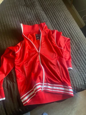 Röd zip hoodie från Jack & Jones - Röd hoodie från Jack & Jones med vit dragkedja och vita detaljer längs muddar och krage. Tillverkad i ett mjukt material med två fickor framtill och ribbade muddar. Perfekt för en sportig och avslappnad stil.