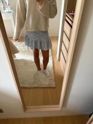 Blå kortkjol med volang och blommor - Söt blå kortkjol med vitt blommönster och volangkant. Kjolen har en omlott-look med knyt i sidan och är perfekt för en fräsch och somrig stil. Lätt och luftig, passar till både stickad tröja och t-shirt.🩷passar mig som brukar ha xs!💕