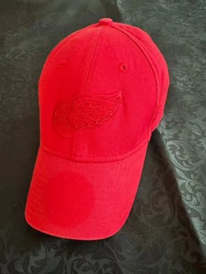 Röd keps NHL  NEW ERA  - Snygg röd keps med böjd skärm och broderad vinge-logga framtill. Kepsen har ventilationshål och justerbar baksida. Tillverkad i bomullstyg och har en klassisk passform. Perfekt för dig som vill ha en stilren och sportig look.