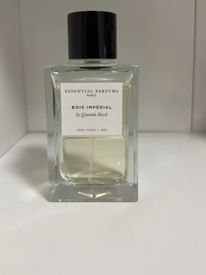 Bois Impérial parfym från Essential Parfums - Bois Impérial från Essential Parfums är en elegant parfym i en stilren, rektangulär glasflaska med svart kork. Doften är skapad av Quentin Bisch och har en ljusgul vätska. Flaskan har en minimalistisk etikett med tydlig text.