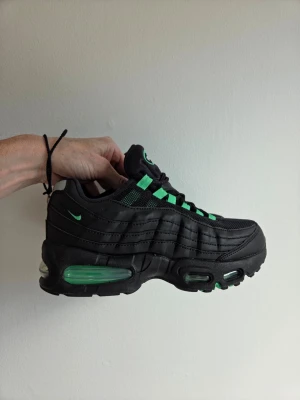 Svarta Nike Air Max 95 med gröna detaljer - Nike Air Max 95 sneakers i svart med gröna detaljer och synlig Air-sula. Skon har en chunky siluett, mesh och läderpaneler samt gröna accenter på snörningen och loggan. Perfekt för dig som gillar streetwear och vill sticka ut. I ny skick.