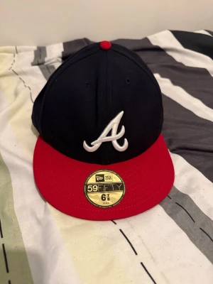 New Era 59FIFTY Atlanta keps - Snygg 59FIFTY keps från New Era med Atlanta-logga framtill. Kepsen har mörkblå krona, röd skärm och röd knapp på toppen. Klassisk flat brim och broderad vit 'A' framtill. Insidan har märkning och MLB hologram. Perfekt för dig som gillar streetwear.