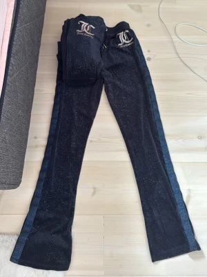 Juicy Couture mörkblått velourset - Snyggt mörkblått velourset från Juicy Couture med glittriga detaljer och blåa ränder längs sidorna. Setet består av byxor med utsvängda ben och matchande zip-hoodie med logga i silver på bröstet och ryggen. Perfekt för chill dagar hemma.