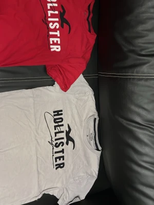 Två Hollister t-shirts i rött och vitt - Säljer två snygga t-shirts från Hollister, en i rött och en i vitt. Båda har klassisk logga och tryck framtill. Sköna och stilrena t-shirts i bomull med normal passform och korta ärmar. Perfekta till jeans eller shorts.