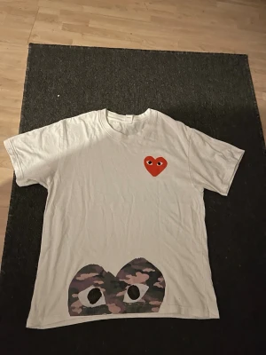 Vit CDG tröja  - Vit cdg tröja med ett rött hjärta på bröstet och ett stort kamouflagemönstrat hjärta längst ner. Klassisk passform och rund hals. Perfekt för dig som gillar streetwear och coola prints.