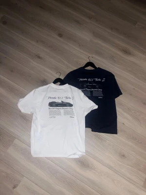 Två t-shirts med Porsche-tryck - Säljer två t-shirts i klassisk modell, en vit och en marinblå, båda med tryck på ryggen och liten text på bröstet. Sköna och stilrena, perfekta till jeans eller shorts. Materialet känns som mjuk bomull och passformen är normal. Båda är i nytt skick. Hör av er vid funderingar.