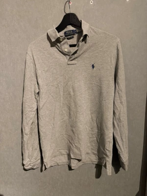 Grå långärmad piké från Polo Ralph Lauren - Klassisk grå långärmad pikétröja från Polo Ralph Lauren med krage, knappar vid halsen och den ikoniska broderade loggan på bröstet. Tillverkad i mjukt bomullsmaterial som är skönt mot huden. Perfekt för en avslappnad men ändå stilren look.