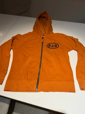 Orange hoodie med dragkedja från Parajumpers  - Snygg orange hoodie från Parajumpers med dragkedja framtill, huva med snörning och två fickor. Stor P.J.S-logga på bröstet och patch på ärmen. Perfekt för dig som vill sticka ut med en färgstark och bekväm hoodie.