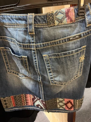 Jeanskjol med etnomönster från Miss Me - Säljer en kort jeanskjol från Miss Me med snygga etno-inspirerade broderier i färgerna röd, rosa, beige och svart längs nederkanten och linningen. Kjolen har klassiska jeansfickor, bälteshällor och kontrastsömmar. Perfekt för dig som vill ha en unik denimlook.