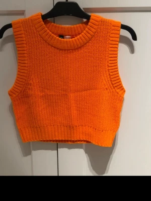 Orange stickad väst från Divided H&M - Säljer en orange stickad väst från Divided H&M. Västen är cropped, har bred ribbad kant vid hals och ärmhål samt en chunky stickad struktur. Perfekt att styla över en skjorta eller t-shirt för en trendig look.