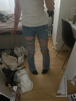 Ljusblå jeans med stora hål - Säljer ett par ljusblå jeans med stora, trendiga hål framtill på båda benen. Jeansen har rak passform och klassisk femficksdesign. Perfekta för dig som gillar en edgy och avslappnad stil. Materialet är denim i bomull.